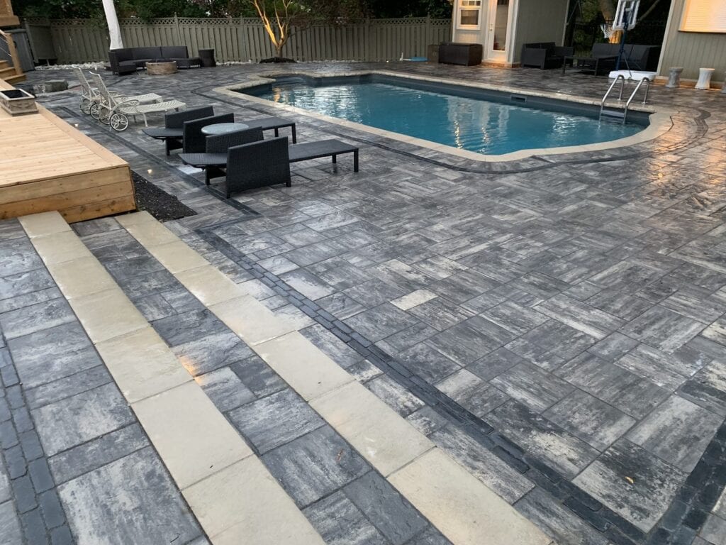 Charcoal and grey interlocking paver pool deck with contrasting cream border bands in a landscaped backyard